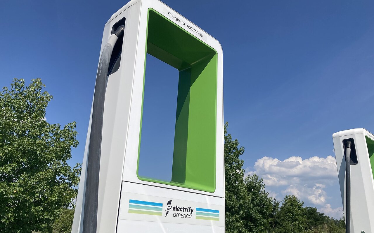 Borne de recharge rapide Electrify America, colonne blanche à arche verte, photographiée en extérieur sous un ciel bleu, avec une seconde borne sur la droite