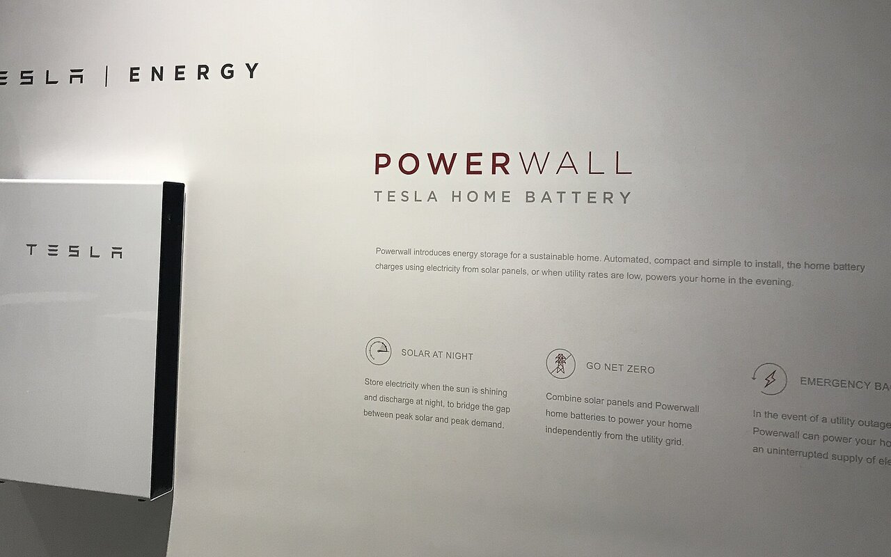 Une batterie domestique Powerwall de Tesla, blanche et verticale, présentée dans un espace d'exposition devant un panneau mural Tesla Energy