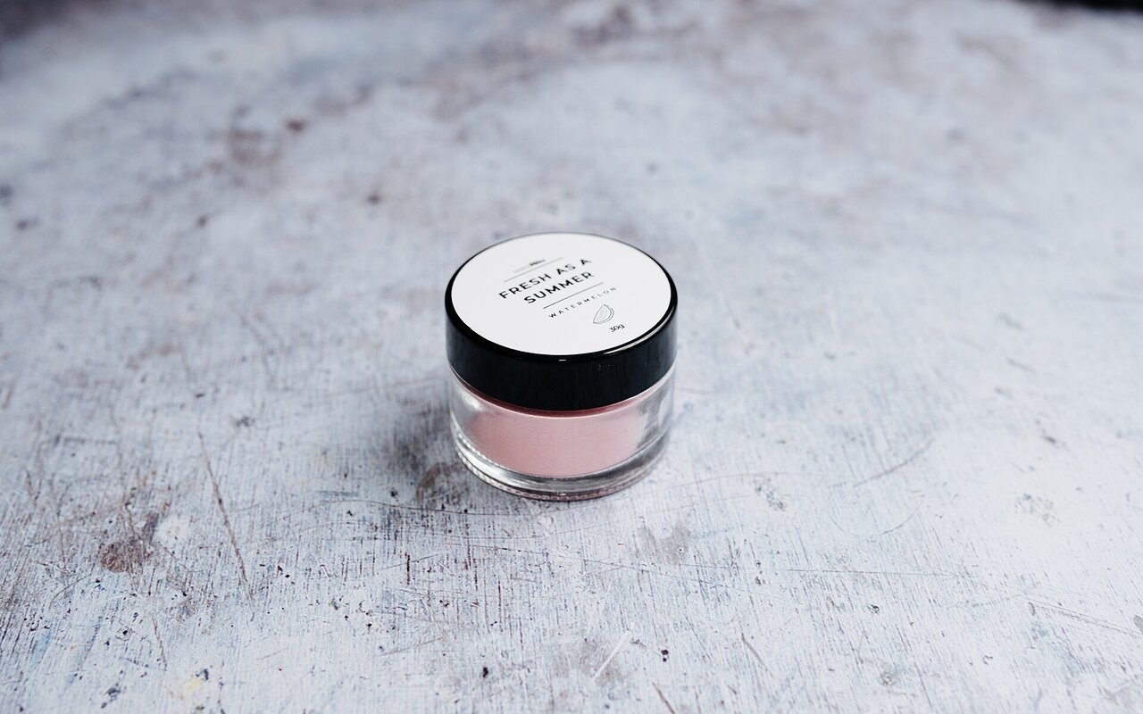 Petit pot en verre contenant un produit cosmétique rose clair sur une surface texturée