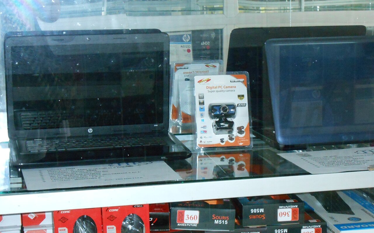 Ordinateurs portables noirs exposés derrière la vitrine d'un magasin d'informatique, au-dessus d'un présentoir d'accessoires et de chargeurs
