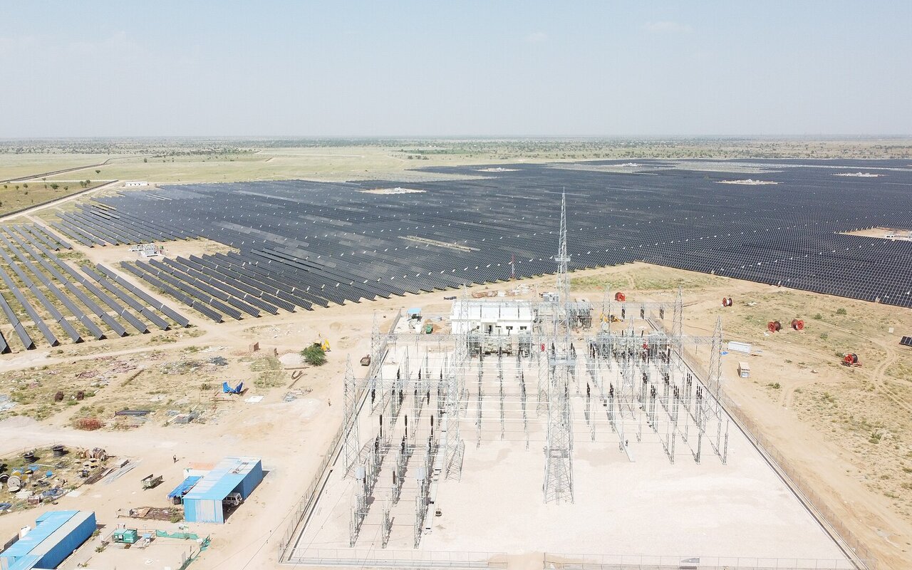 Vue aérienne d'une vaste centrale solaire photovoltaïque au sol, avec au premier plan un poste électrique de raccordement et ses pylônes.