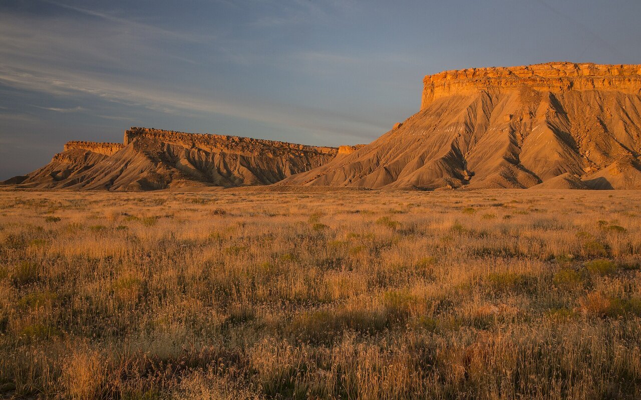 Paysage de terres publiques de l'Utah : une mesa aux falaises ocres éclairée par la lumière rasante du couchant domine une vaste étendue plate d'herbes sèches.