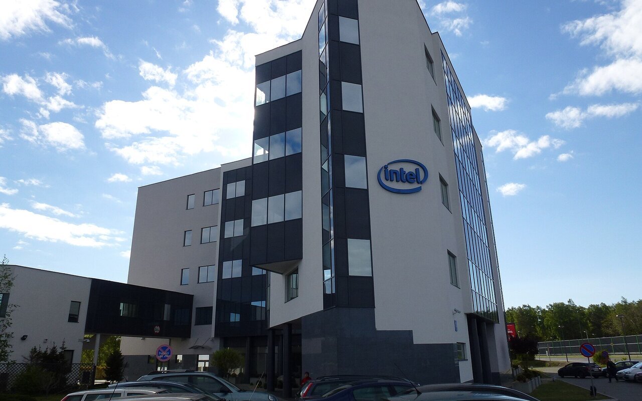 Immeuble de bureaux moderne portant le logo Intel sur sa façade, sous un ciel bleu, avec des voitures stationnées au premier plan