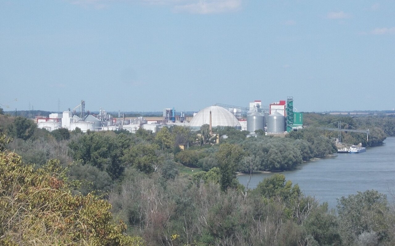 Usine d'éthanol au bord d'un fleuve, avec ses silos, ses cuves cylindriques et ses dômes de stockage, vue depuis la rive opposée boisée