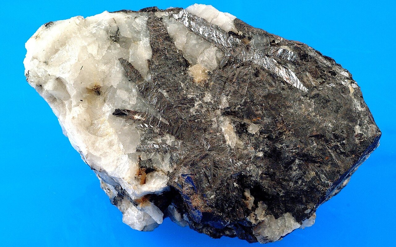 Échantillon minéral de wolframite, minerai de tungstène sombre et métallique, associé à du quartz blanc, photographié sur un fond bleu.