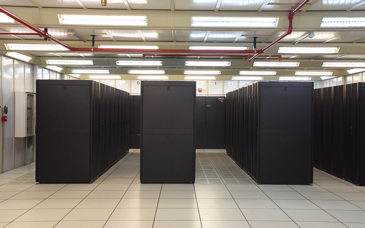 Salle d'un centre de données : rangées de baies de serveurs noires fermées, posées sur un plancher technique clair, sous un plafond de luminaires.