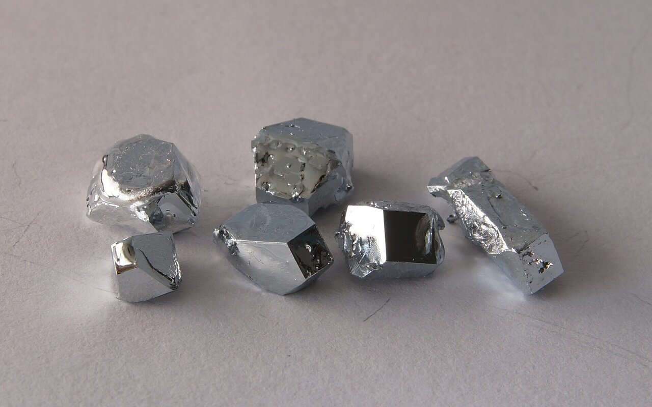 Plusieurs cristaux de gallium métallique gris et brillants, aux faces anguleuses, posés sur une surface claire