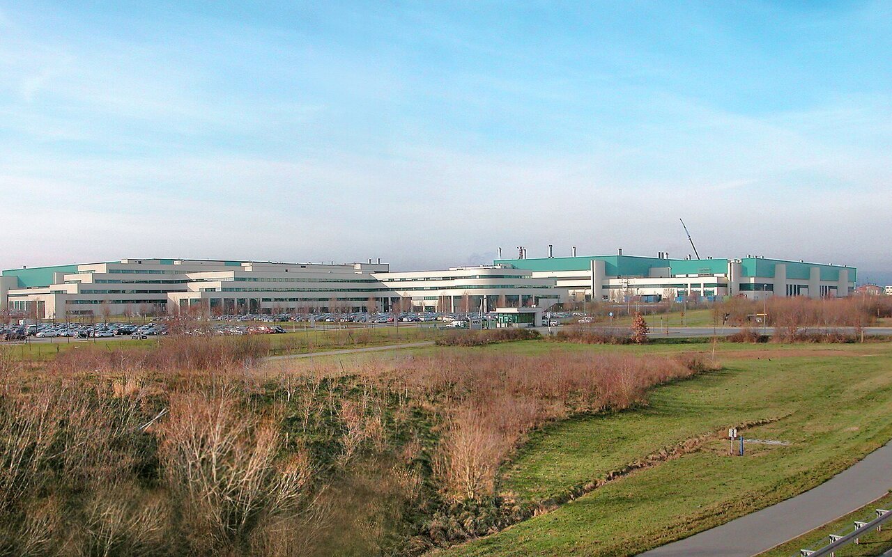 Vue d'ensemble de l'usine de semi-conducteurs de Dresde-Wilschdorf, longs bâtiments industriels blancs et vert d'eau entourés de champs et d'un parking.